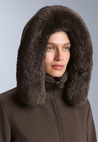 Hnedý parka s hrubou, umelou kožušinou na kapucni. Kapucňa má nadýchanú textúru, rámujúcu tvár a kontrastuje so hladkým materiálom bundy.