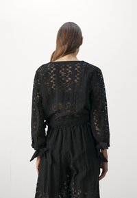 Musta läpinäkyvä paita, jossa on rakenteellinen kuvio ja pitkät hihat. Varustettu sidottavalla vyötärödetaililla ja suoralinjaisella kaula-aukolla.