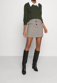 Femme portant un pull à col en V vert foncé sur une chemise blanche à col, une mini-jupe à boutons motif pied-de-poule, et des bottes noires montantes jusqu'aux genoux.