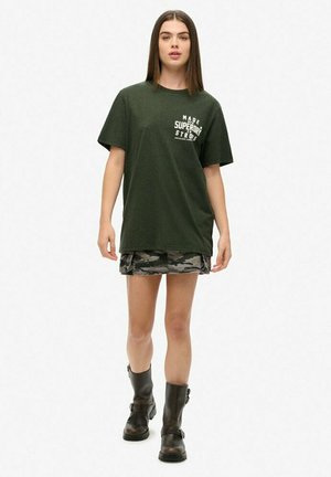Camiseta de gran tamaño de color verde oscuro con texto gráfico blanco, combinada con una falda mini de camuflaje y terminada con botas negras hasta el tobillo.