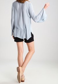 Femme portant un chemisier long à manches bleu clair, un short noir et des chaussures à talons carrés beiges, marchant sur un fond léger uni.