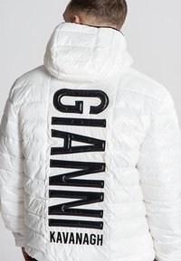 Bijela jakna s podstavom koja ima kapuljaču i velike crne natpise "GIANNI KAVANAGH" na leđima, sa quilted teksturom i sjajnim naglascima.
