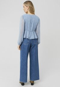 Blouse florale bleu clair avec des manches longues en voile et un design peplum, associée à un jean bleu taille haute à jambes larges et des chaussures à talons noires.