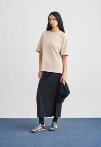 Beżowy bawełniany oversize t-shirt, czarna midi spódnica z bocznym rozcięciem, srebrne buty sportowe i czapka w czarnym kolorze, na jasnym tle.