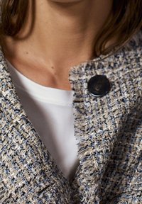 Kurtka z materiału tweed w kremowych, niebieskich i szarych odcieniach z postrzępionym kołnierzem i dużym czarnym guzikiem. Noszona na gładkiej białej bluzce.