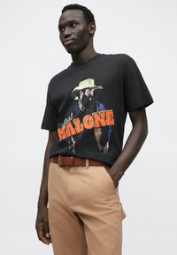 Tricou negru din bumbac, cu un imprimeu grafic al unui bărbat cu barbă și pălărie de cowboy, cu text portocaliu "Post Malone". Asociat cu pantaloni bej.