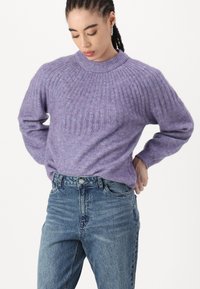 Pull en tricot violet avec une texture côtelée et un col haut, associé à un jean taille haute bleu clair. Matériau doux et confortable.
