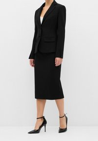 Femme portant un blazer noir cintré, une jupe crayon assortie longueur genoux, un haut blanc et des chaussures noires à talons hauts avec bride à la cheville.