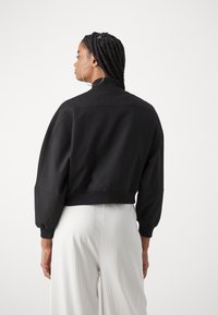Veste noire courte avec un col montant, des épaules tombantes et des poignets côtelés. Tissu lisse au design épuré ; portée sur un pantalon de couleur claire.