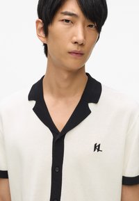 Fehér kötött polo póló fekete gallérral és díszítéssel, fekete logóval a bal oldalon és gombos elülső résszel. Sima tapintású.