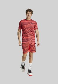 Conjunto deportivo rojo que incluye una camiseta de manga corta texturizada con patrones ondulados y pantalones cortos a juego, combinado con zapatillas deportivas blancas y negras.