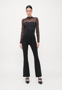 Černý krajkový bodysuit s dlouhými rukávy spárovaný s form-fitting černými rozšířenými kalhotami a lodičkami na jehlovém podpatku s přezkovými akcenty. Hladká textura a elegantní silueta.