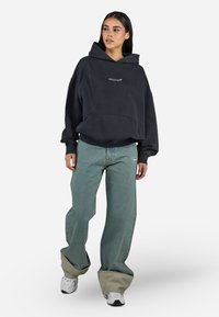 Zwarte oversized hoodie met een voork pocket en logo, gecombineerd met lichtblauwe wijde jeans met een logo, en witte sneakers.