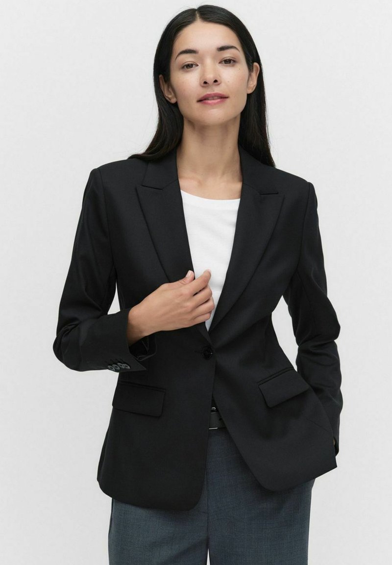 Svart skräddarsydd blazer i släthet, med enstaka knappstängning och två framfickor. Bärs över en vit topp, strukturerad passform.