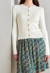 Cardigan bianco a coste con bottoni dorati, abbinato a una gonna a motivi in turchese e marrone con un design fluido e pieghe.