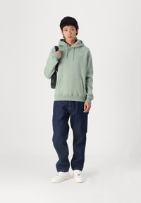 Sudadera verde clara con un bolsillo delantero y capucha con cordón, combinada con jeans sueltos de un azul oscuro y zapatillas blancas. Bolso de hombro negro.