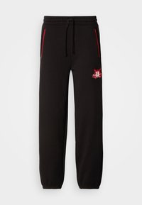 Zwarte sportbroek met een elastische tailleband, rode accent ritsen en een opvallend geborduurd logo op de linkerdij. Gladde textuur, tapersnit.