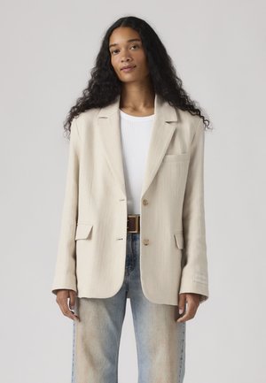 Femeie cu păr lung și creț, purtând un sacou bej peste o cămașă albă și blugi albaștri decolorați cu o centură maro, stând în fața unui fundal simplu.