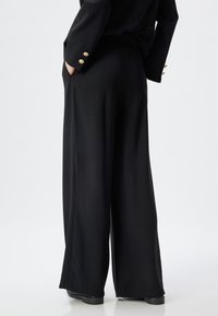 Pantalon large noir avec une texture fluide, doté d'une taille élastique et d'une silhouette élégante, associé à un blazer noir ajusté.