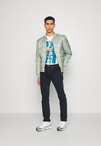 Cazadora bomber acolchada de color verde claro, camiseta gráfica blanca, jeans de mezclilla oscura y zapatillas deportivas grises con acentos en neón.