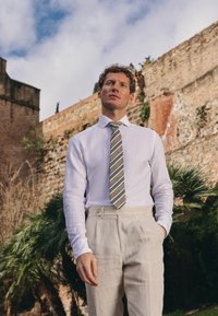 Witte lange mouwen shirt gecombineerd met een grijze gestreepte stropdas. Lichtbeige broek met zijzakken, gladde stof, tegen een gestructureerde muur.