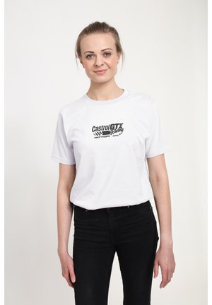 Uśmiechnięta kobieta o jasnych włosach, ubrana w białą koszulkę Castrol GTX Racing oraz czarne spodnie, stojąca na jednolitym tle.