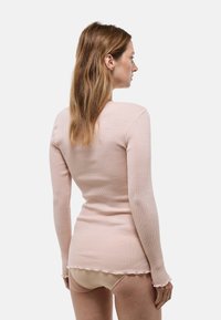 Femme aux longs cheveux châtain clair portant un haut ajusté à manches longues rose pâle et une lingerie couleur chair, vue de dos sur un fond uni.