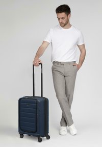 Marineblaue Hartschalenreisetasche mit strukturierten Streifen, zwei schwarzen Teleskopgriffen und vier Rädern, die neben einer Person in hellen Hosen steht.