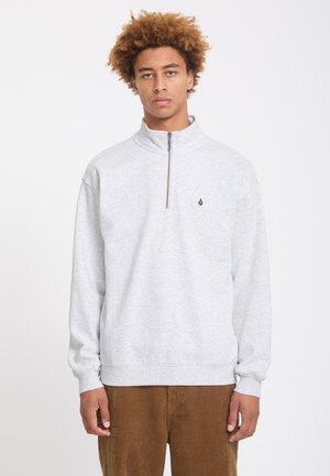 Jeune homme aux cheveux bouclés portant un sweat-shirt gris clair à fermeture éclair quart et un pantalon en velours côtelé marron, debout devant un fond blanc uni.