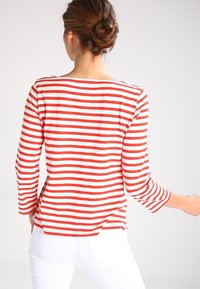 Rot-weiß gestreiftes Langarmshirt mit Bootsausschnitt und seitlichen Schlitzen, aus einem weichen Stoff gefertigt. Die Rückansicht zeigt die Stylingdetails.
