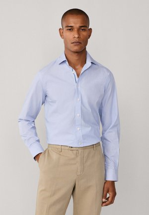 Hombre con camisa de botones azul claro metida dentro de unos pantalones beige, de pie con una mano en el bolsillo frente a un fondo liso.