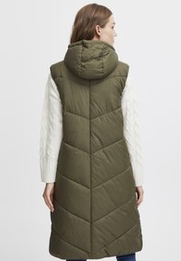 Olivgrön puffer väst med huva, med chevron-quilting och lång längd. Vesten är matchad med en vit kabelstickad tröja.
