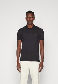 Camisa polo negra de manga corta hecha de algodón, con cuello, cierre de tres botones y un pequeño logotipo en el lado izquierdo del pecho.