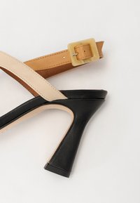 Høyhælte sandaler med svart lærsåle, beige og brune remmer, og en firkantet smaragdspenne som accent. Glatt tekstur med minimal søm.