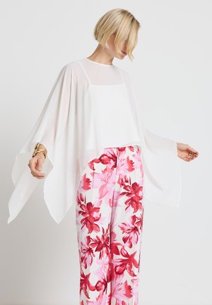 Frau mit kurzen blonden Haaren, die eine durchsichtige weiße, fließende Bluse und weiße Hosen mit großem rotem und rosa Blumenmuster trägt, steht mit ausgestreckten Armen.