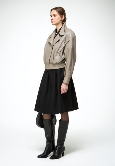 Femme debout portant une veste en cuir marron clair, une jupe noire jusqu'aux genoux, des bottes noires à talons hauts et tenant un sac à main noir.