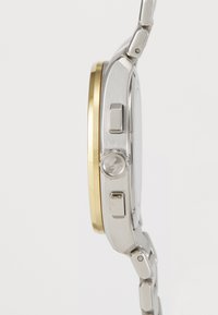 Orologio in metallo argento e oro con cassa rotonda, pulsante e maglie del bracciale testurizzate. Presenta una superficie spazzolata e dettagli lucidi.