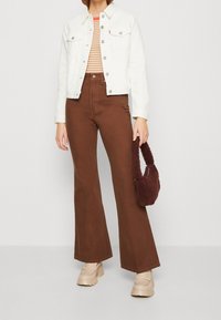 Weiße Jeansjacke, gestreiftes Oberteil, braune Schlaghose, beigefarbene Plateauschuhe und eine braune Fleece-Handtasche. Fokus auf Textur und Farbe.