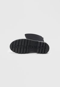 Semelle de botte en caoutchouc noir avec un motif de semelle profonde, présentant une texture lisse et un nom de marque en relief, mettant en avant la durabilité et l'adhérence.