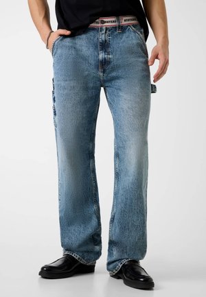 Hombre con vaqueros rectos azul claro con cinturilla visible roja y blanca con la palabra "HYSTERIC", zapatos negros y camisa negra con las manos en los bolsillos.