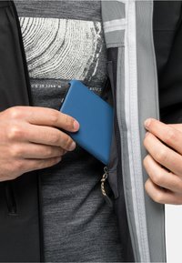 Ein blauer Handy wird in eine mit Reißverschluss ausgestattete Tasche einer grau-schwarzen Jacke gesteckt. Die Jacke hat reflektierende Akzente und ein Netzfutter.