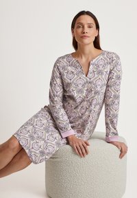 Langärmliges Nachthemd in hellem Lila mit einem komplexen Paisleymuster; verfügt über einen V-Ausschnitt und weiche, pinkfarbene Manschetten. Wird im Sitzen auf einem strukturierten Pouf getragen.
