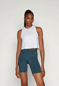 Kobieta o ciemnych włosach związanych w kucyk, ubrana w białą bezrękawnikową bluzkę i ciemnozielone szorty rowerowe Nike, na jednolitym tle.