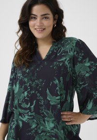 Tunique marine avec motif floral vert, décolleté en V et manches amples. Fait de tissu doux avec une texture lisse.