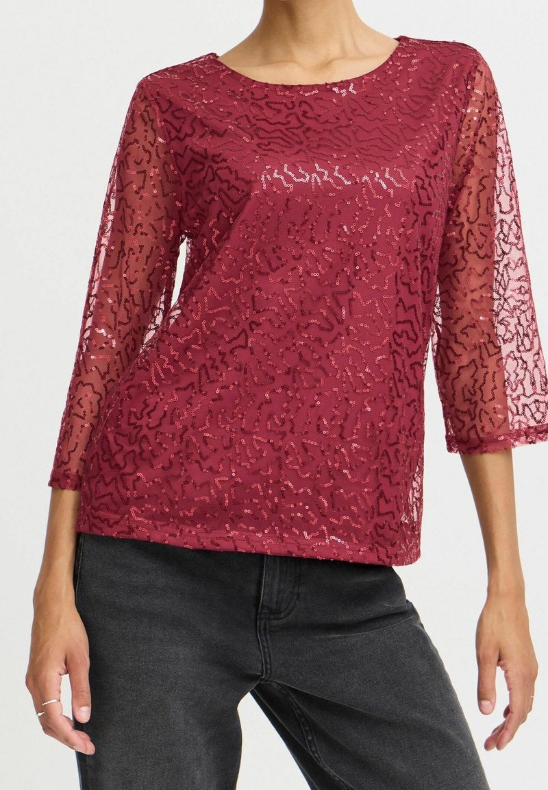 Burgundowy top z cekinami i półprzezroczystymi rękawami o długości trzy czwarte, noszony z czarnymi dżinsami, ukazujący górną część tułowia i ramiona osoby.