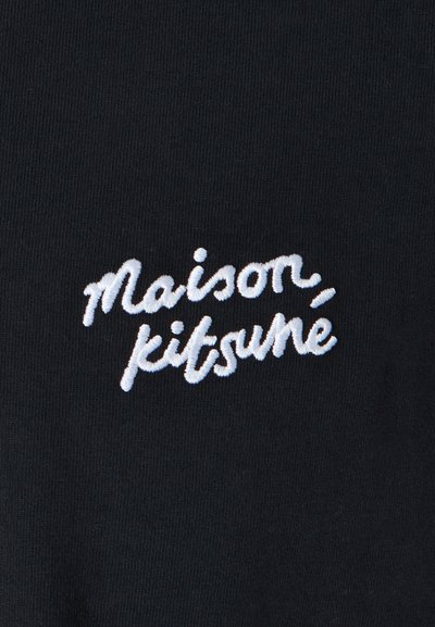 Black fabric featuring white embroidered text reading "maison kitsuné" in a casual, cursive style. Close-up detail of stitching.