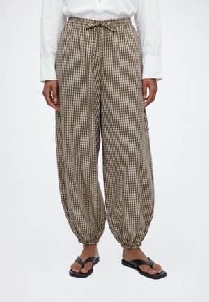 Person iført løstsiddende, brune og hvide ternede bukser med elastiske manchetter og bundet talje, kombineret med sorte thong-sandaler og en hvid skjorte.