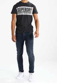 Šedé tričko s bílým logem "SUPERDRY SURPLUS GOODS", spárované s tmavě modrými džiňsy a bílými teniskami. Jednoduchý, neformální outfit.