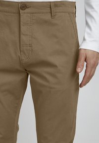 Beige bomull chinos med knappstängning, sidofickor och en jämn textur. Hand placerad vid midjan för skala.