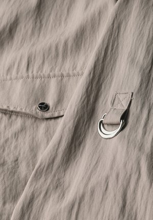 Beige fabric close-up with a textured surface, featuring a black snap button and a silver metal ring attached with a fabric strap.
